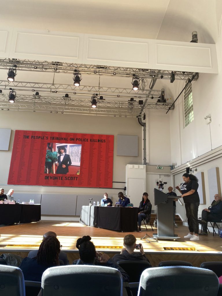 A speaker in an End Police Pursuits t-shirt addresses the People's Tribunal on Police Killings from a lectern. Behind her a large screen shows a portrait of Devonte Scott, set against a red background filled with the names of people who have died following police contact. A panel sits at tables to one side, with an audience watching.
