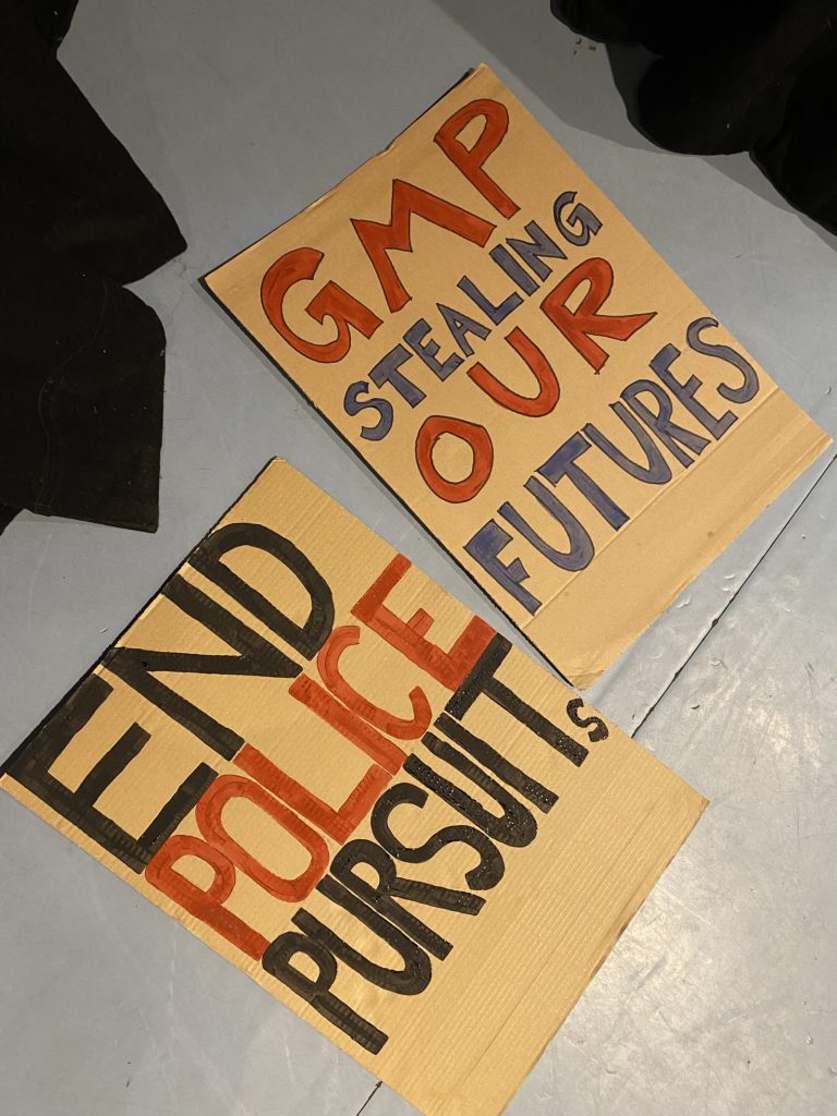 Two hand-painted cardboard placards lying on the ground. One reads "GMP stealing our futures", the other "End police pursuits".