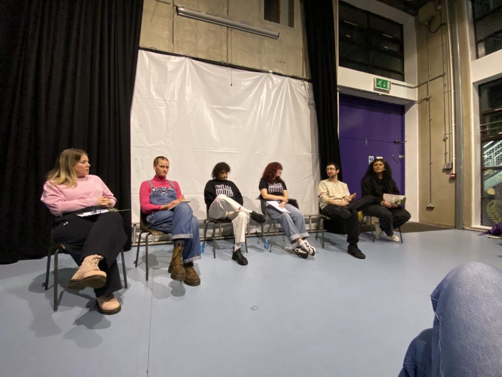 Six speakers seated in a row on a stage at a public meeting, several wearing campaign t-shirts, some holding notes and papers.